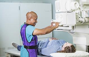 Older bald black man setting up a machine to x-ray a white woman with brown hair laying on a table